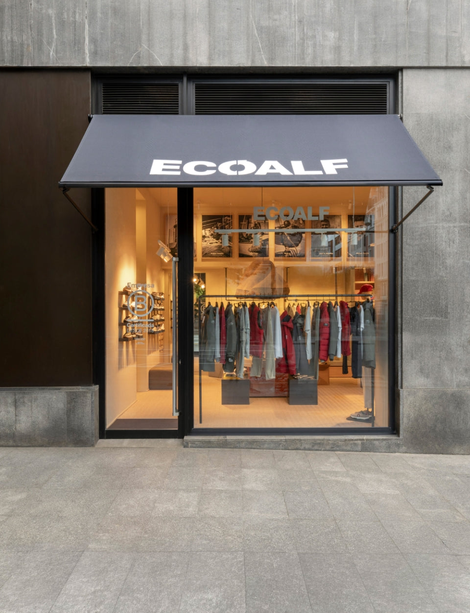 Exterior de una tienda Ecoalf con toldo negro; por el escaparate se ve ropa colgada, suelo de madera y una decoración minimalista.