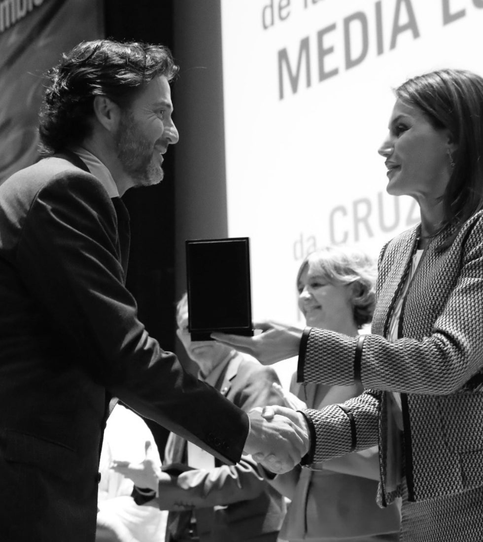 Una persona entrega un premio a otra y se dan la mano en un escenario, mientras al fondo alguien observa la escena.