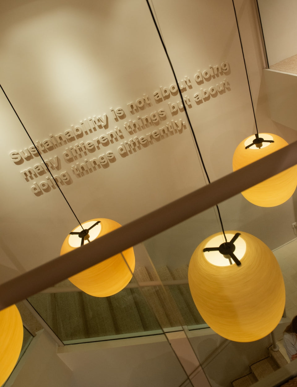 Unas lámparas esféricas de luz cálida iluminan una escalera de diseño moderno, con una frase sobre sostenibilidad en relieve en la pared.