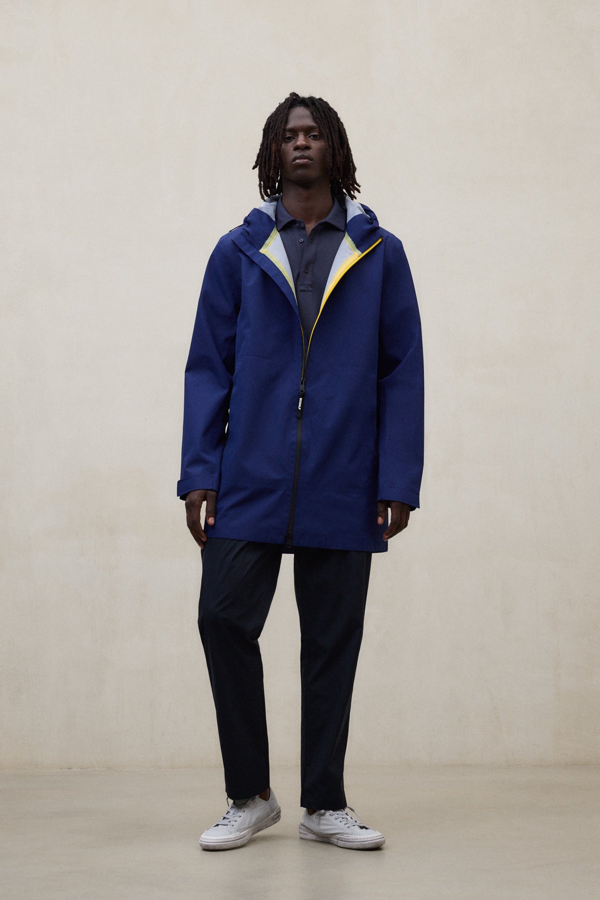 Un hombre en una sala color crema con una chaqueta de Ecoalf azul con detalles amarillos, pantalón negro y zapatillas blancas.
