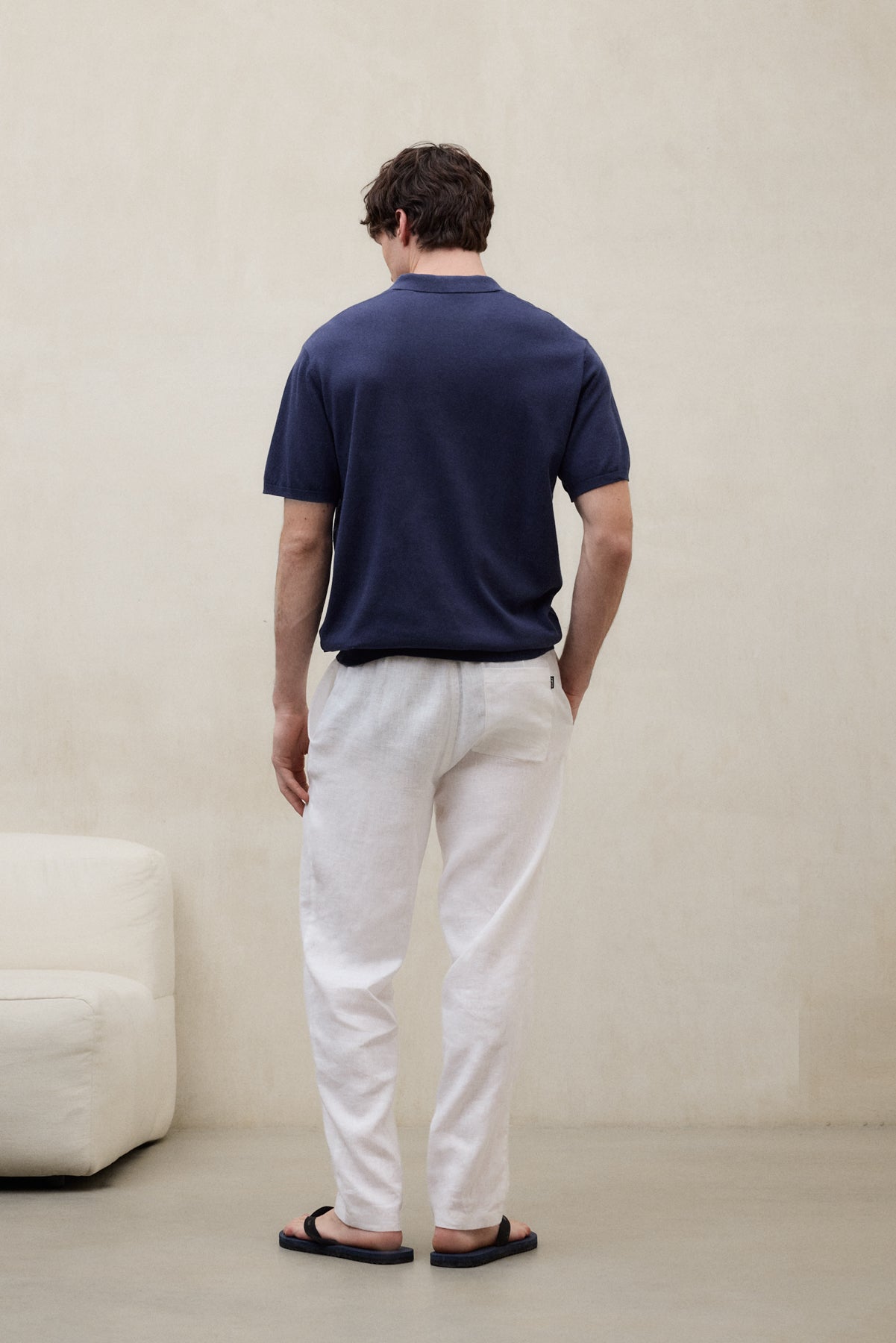 Un hombre de espaldas con una camiseta de Ecoalf azul oscuro y pantalón blanco y sandalias negras, en una sala clara con un sofá blanco a un lado.