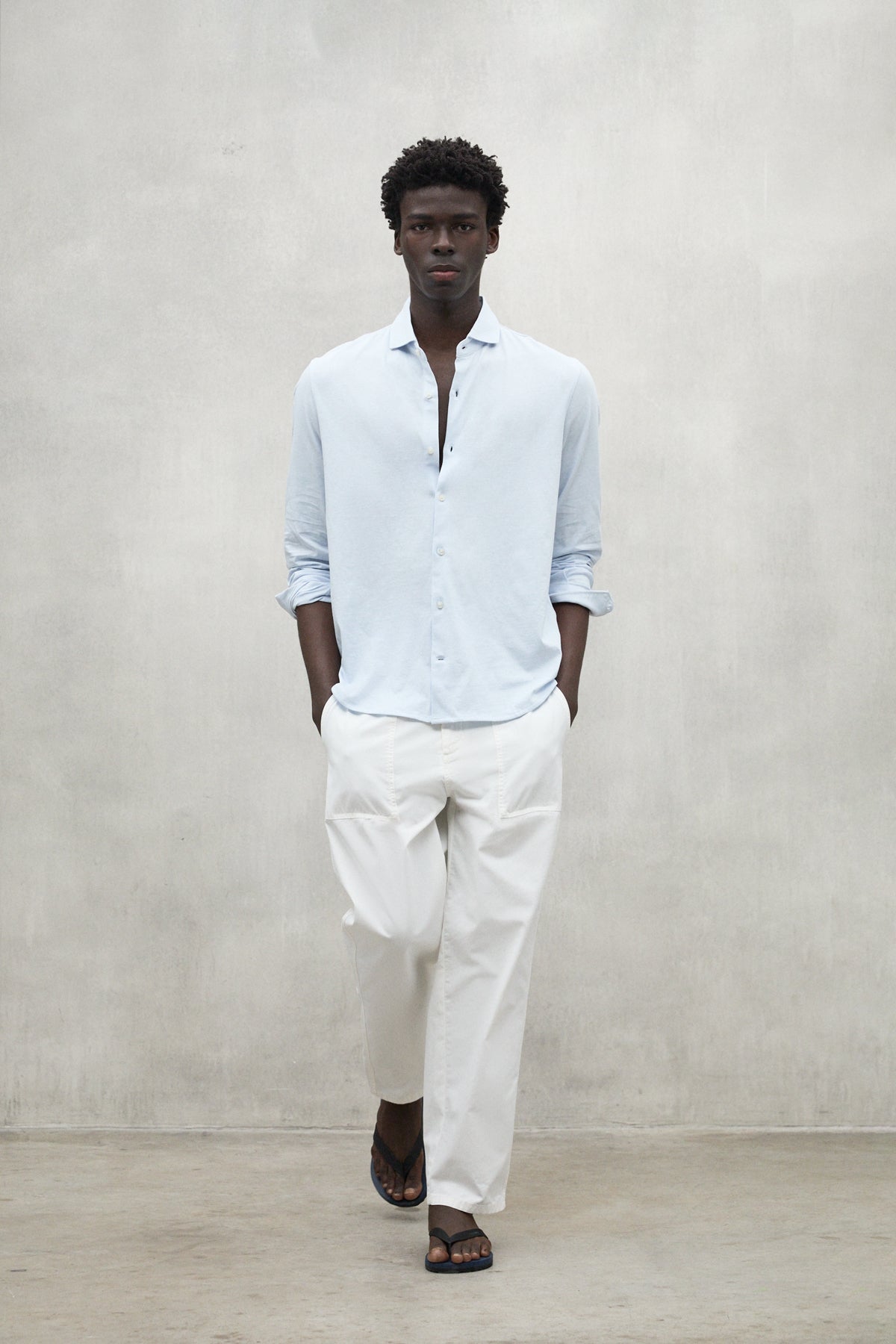 Un hombre con camisa de Ecoalf azul claro, pantalón blanco y sandalias negras camina hacia la cámara sobre un fondo gris neutro.