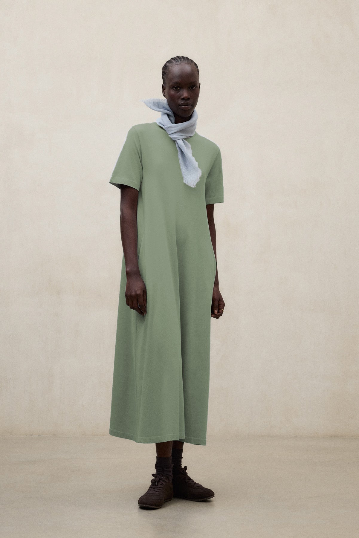 Una mujer en una sala beige con un vestido de Ecoalf largo verde oliva y un pañuelo ligero gris claro, con calcetines a juego y zapatillas oscuras.