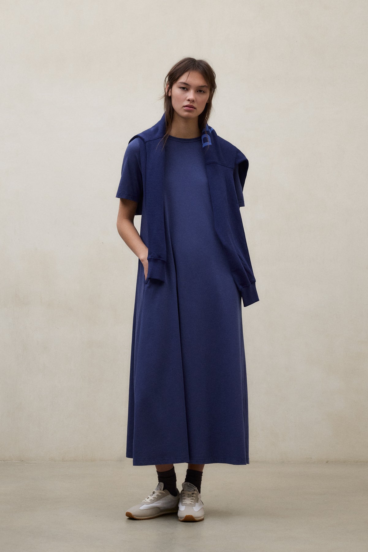 Una mujer con un vestido de Ecoalf largo y suelto azul oscuro y una chaqueta a juego sobre los hombros, con calcetines oscuros y zapatillas blancas, sobre fondo beige.