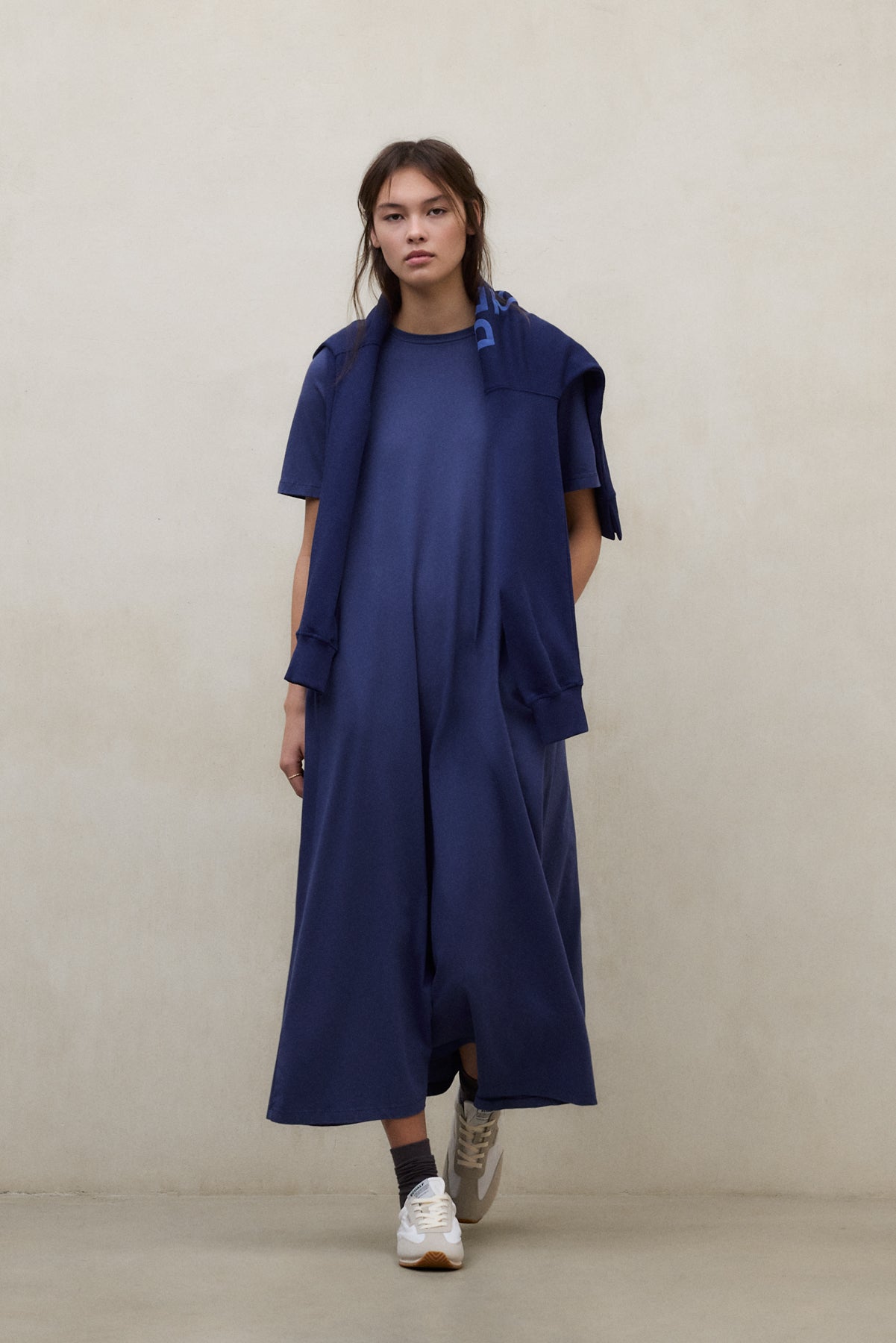 Una mujer con un mono de Ecoalf largo azul marino y una sudadera a juego atada a los hombros, con zapatillas blancas y calcetines oscuros, sobre una pared blanca.