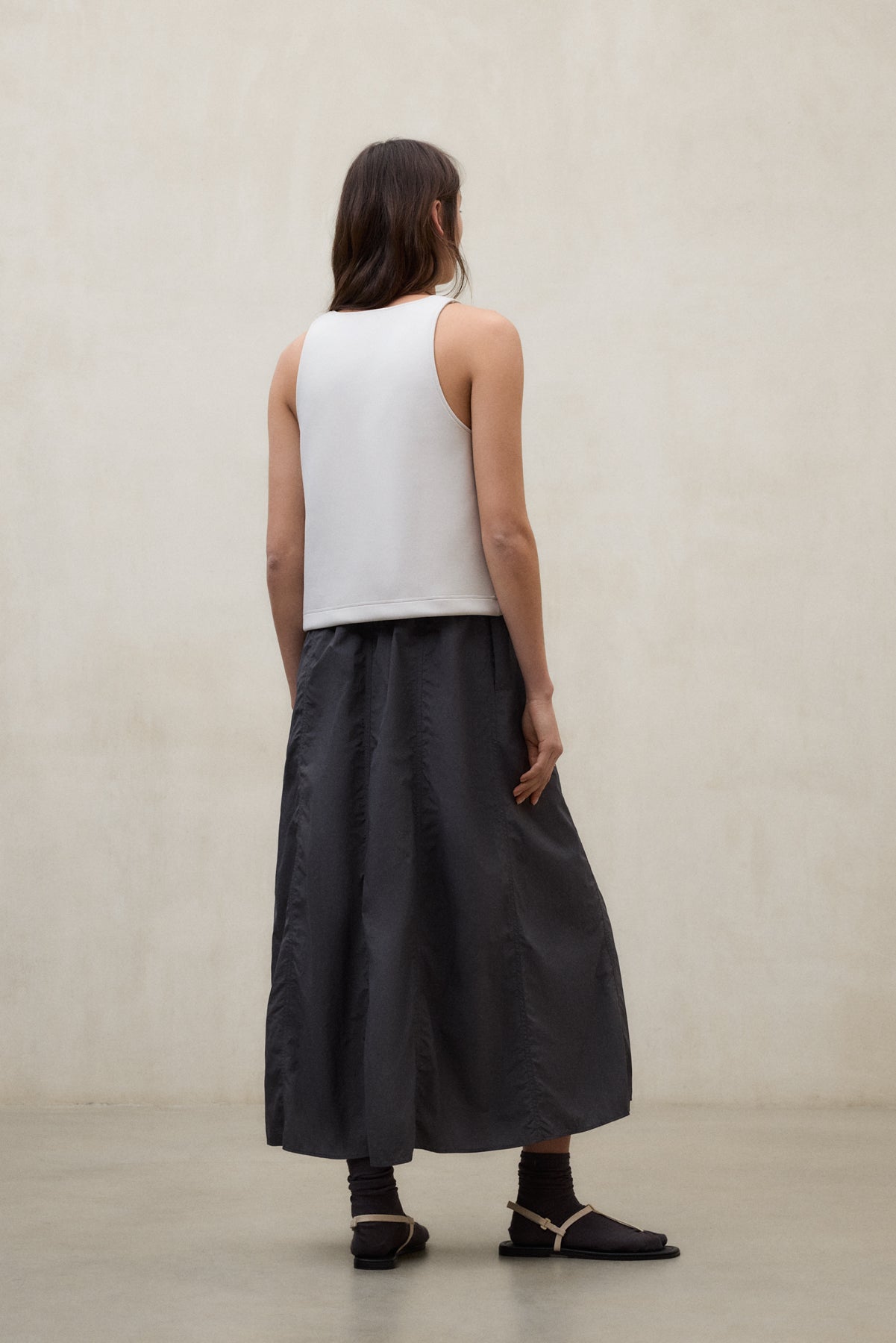 Una mujer de espaldas con una vestido de Ecoalf sin mangas blanco, calcetines oscuros y sandalias claras, sobre una pared beige.