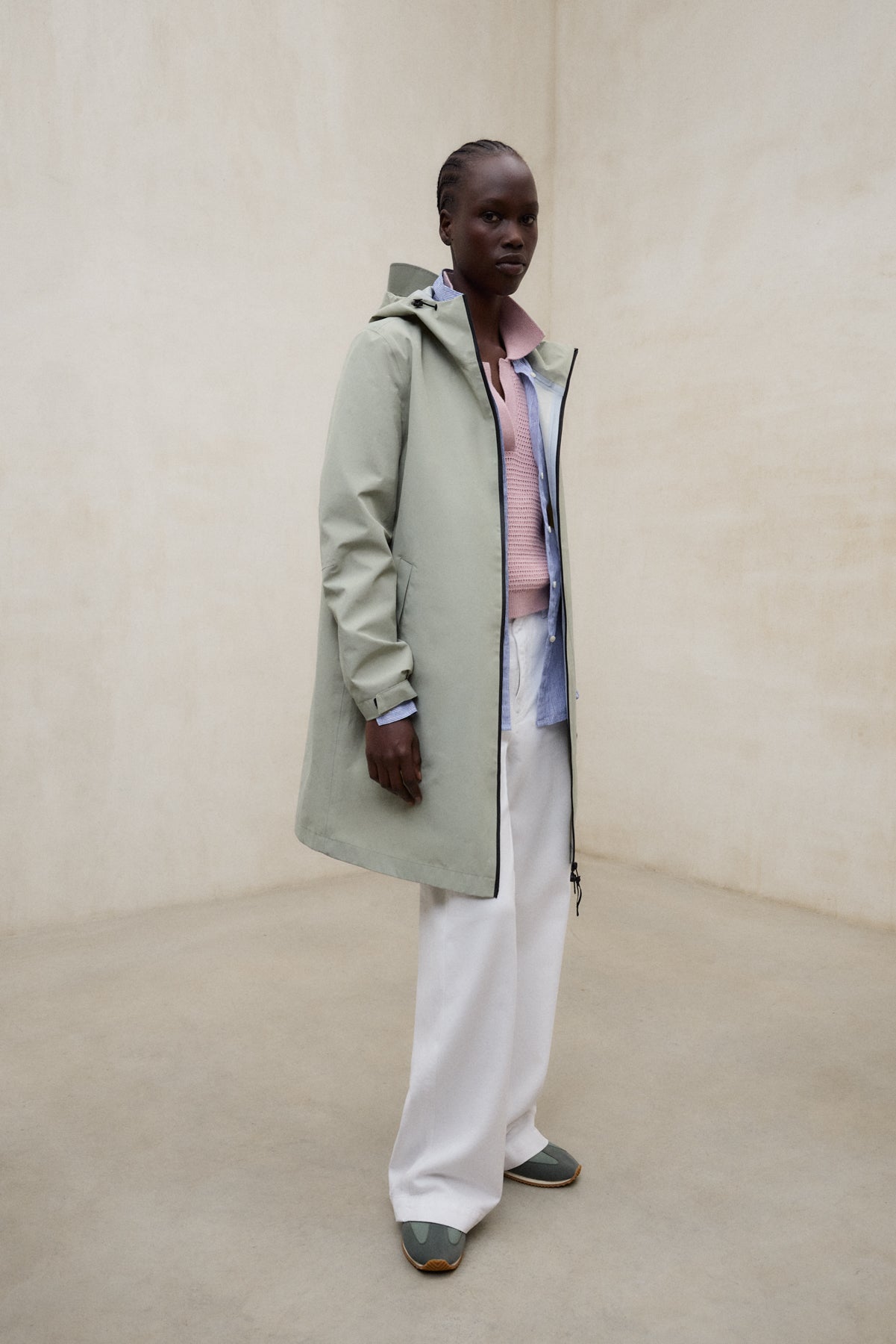 Una mujer en una sala minimalista beige con un abrigo de Ecoalf verde claro con capucha, un jersey rosa claro y pantalón blanco, con zapatillas verdes de suela marrón.
