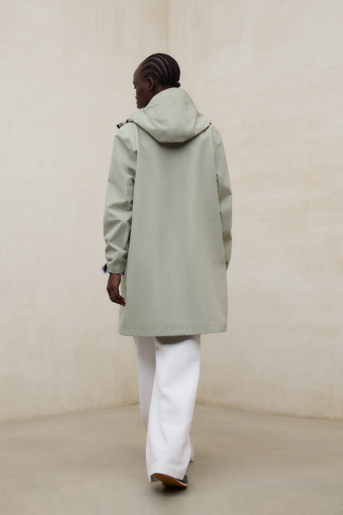 Una persona de espaldas con un abrigo de Ecoalf verde claro con capucha y pantalón blanco, en un entorno minimalista de paredes beige.