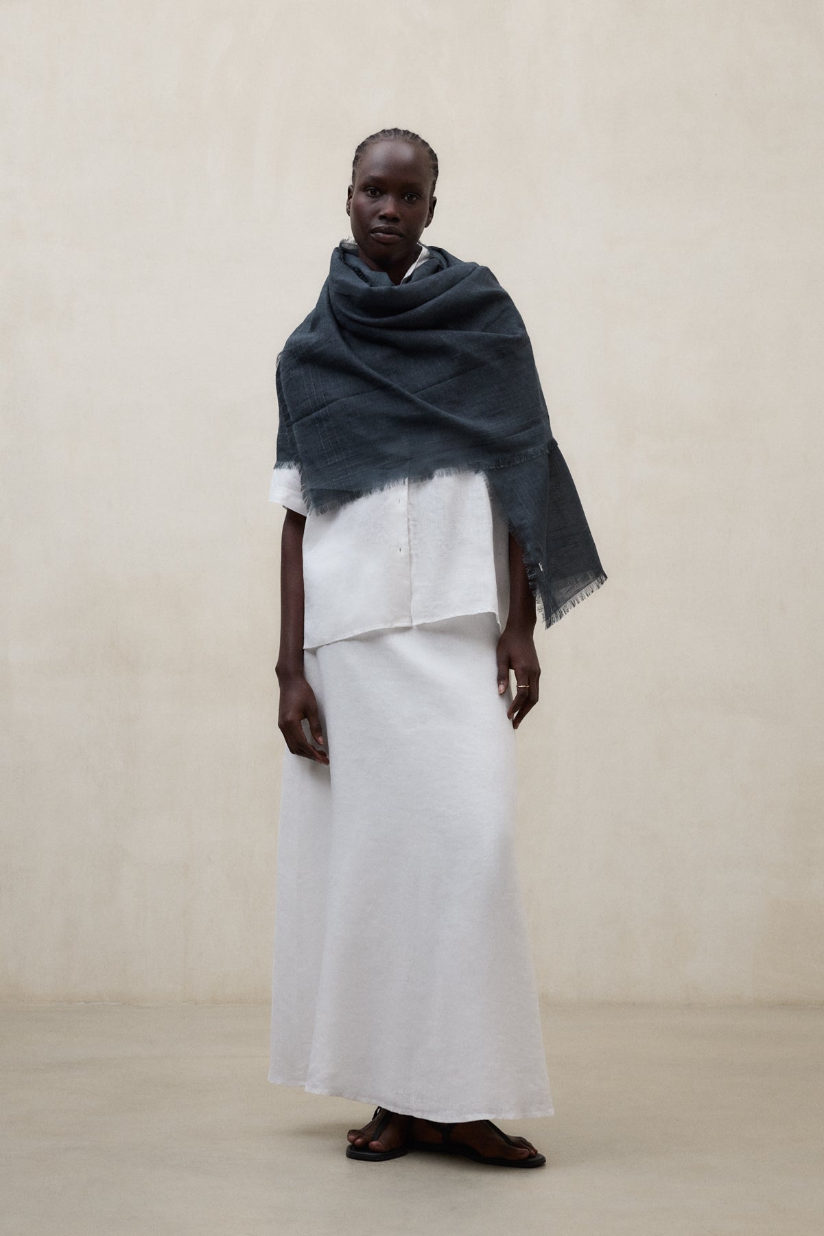 Una mujer con un conjunto de Ecoalf blanco de blusa y falda larga, un chal oscuro sobre los hombros y sandalias negras, sobre fondo neutro.