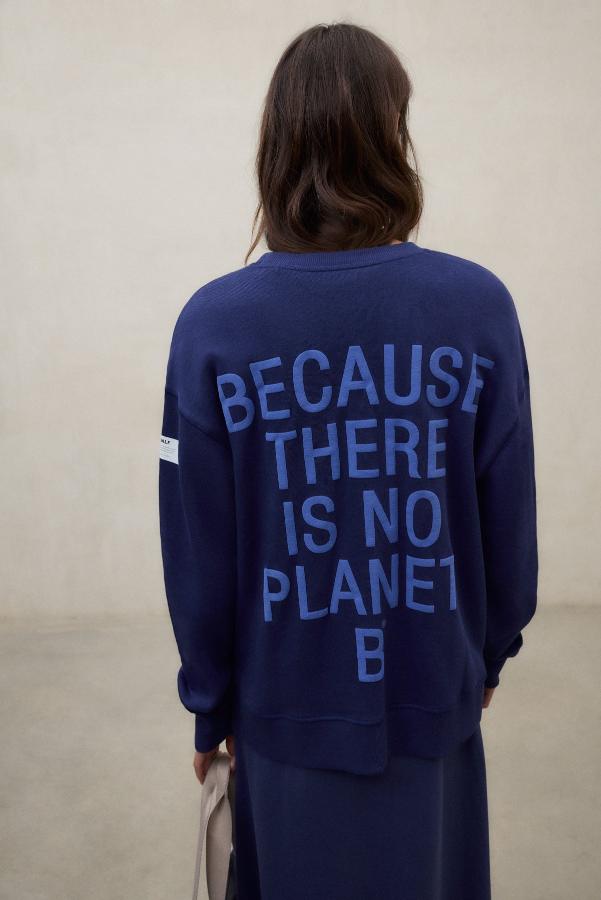 Una mujer de espaldas con sudadera azul de Ecoalf con el lema «Because There Is No Planet B», falda a juego y bolso en la mano.