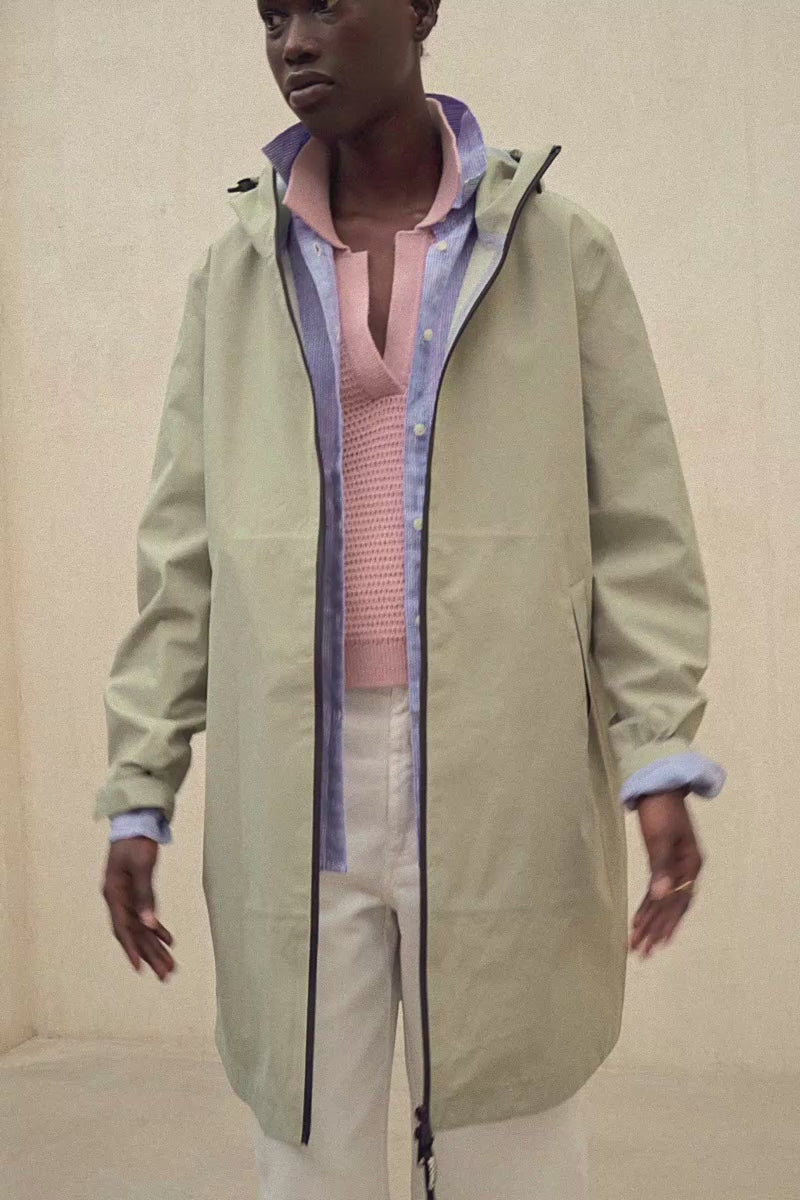 Un modelo con un abrigo de Ecoalf verde claro con capucha sobre una camisa azul y un jersey de punto rosa, con pantalón claro, sobre fondo neutro.